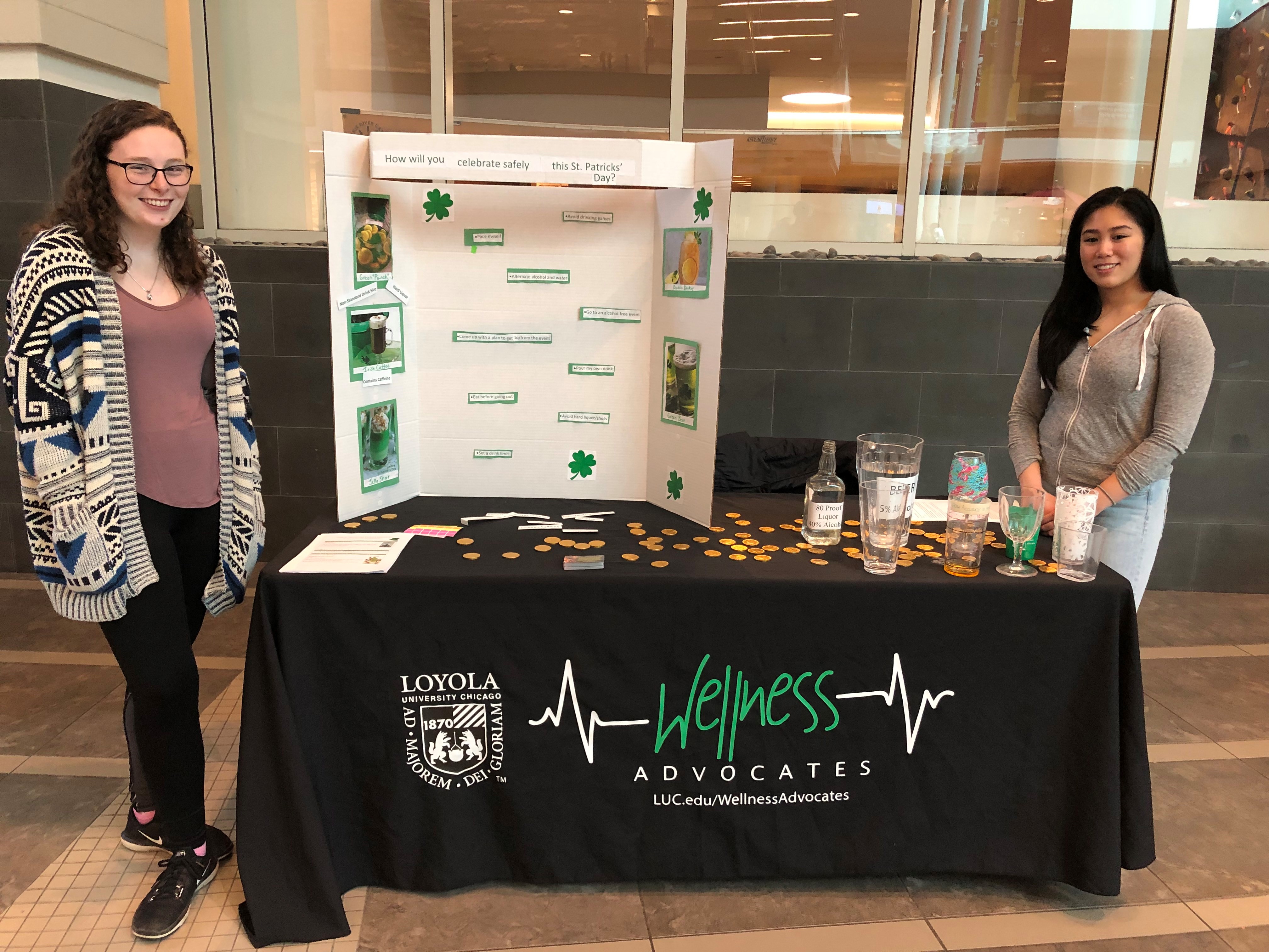 Two students pose near a table decorated for St. Patrick's Day. There is a poster board that reads 